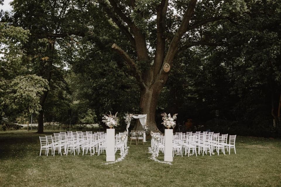 Trauung einer Boho-Hochzeit auf einer Wiese mit einem Traubogen und weißen Stuehlen